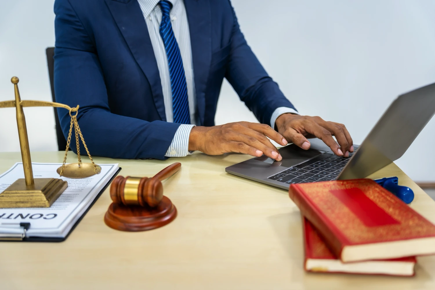 A lawyer typing at his computer at his desk that also has a gavel, a scale sitting on top of a contract. and some books on the table