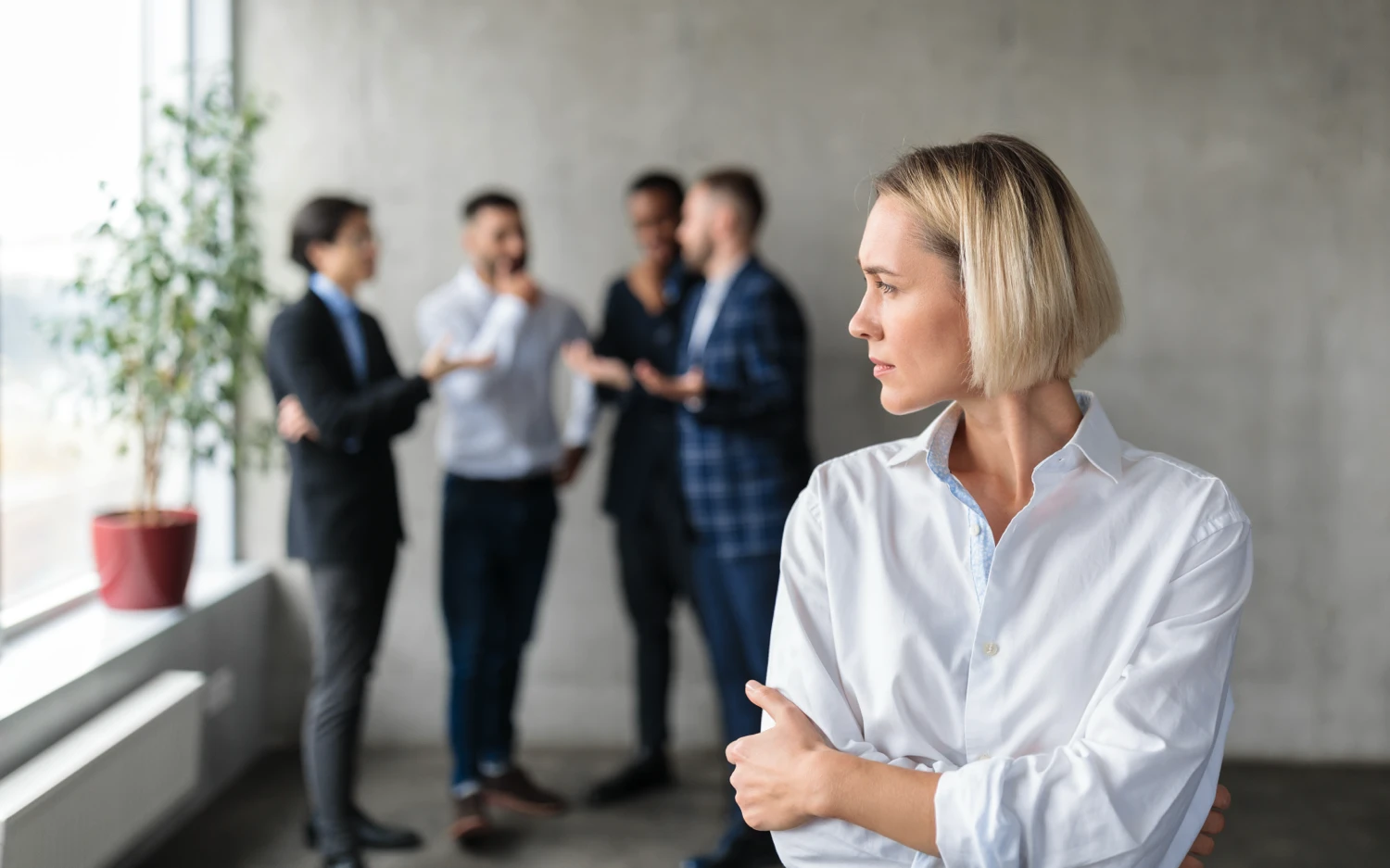 A woman looking sad in an office while her male coworkers talk behind her
