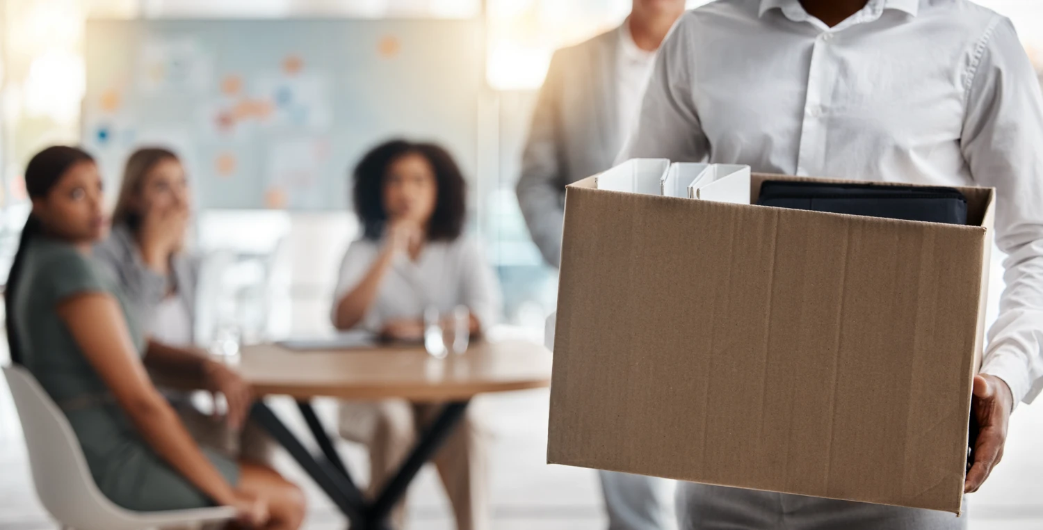 A man carrying a box outside of an office while a male is standing and three female employees are sitting at a table and watching in the background